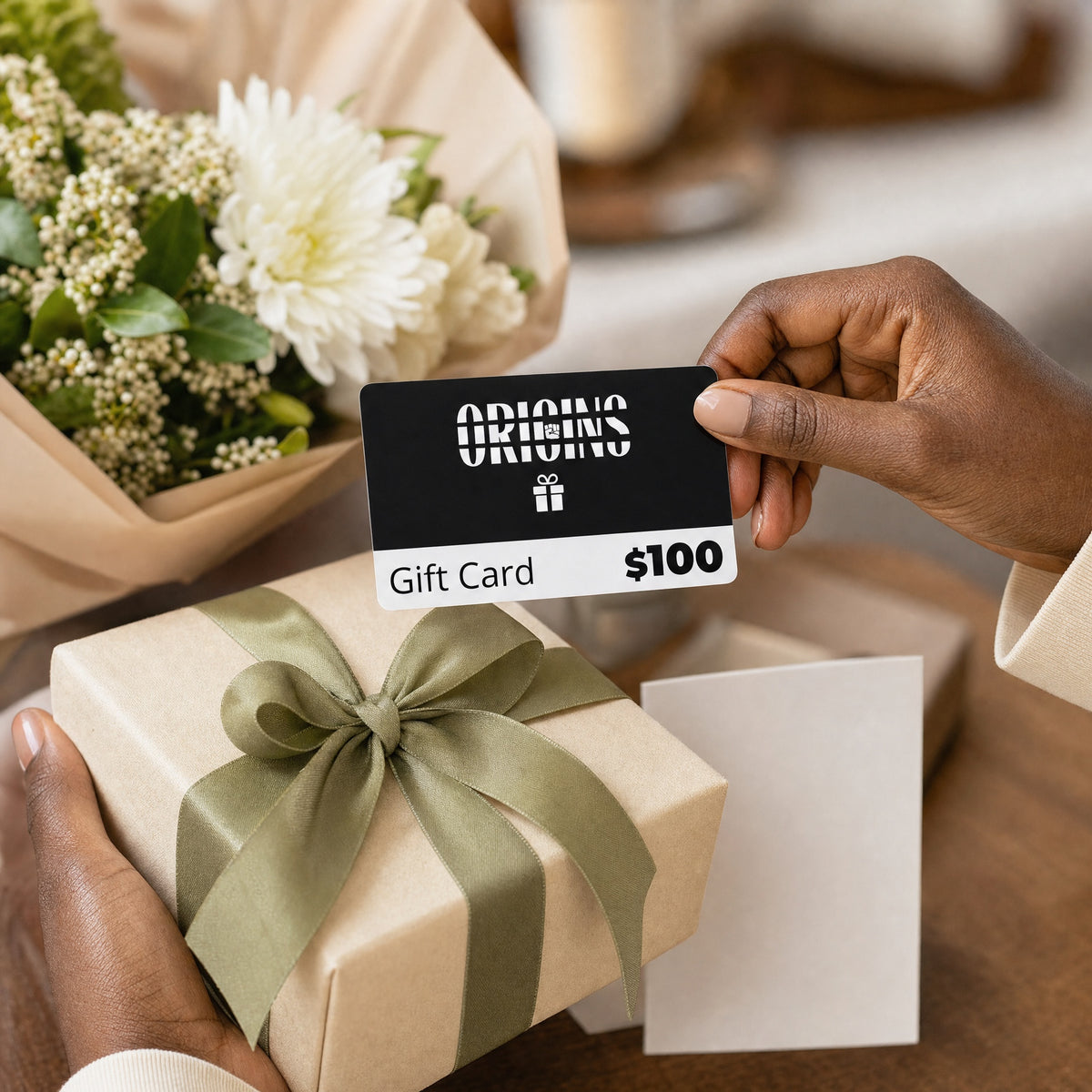 Origins one hundred dollar gift card held above a wrapped gift and flowers in a home setting.